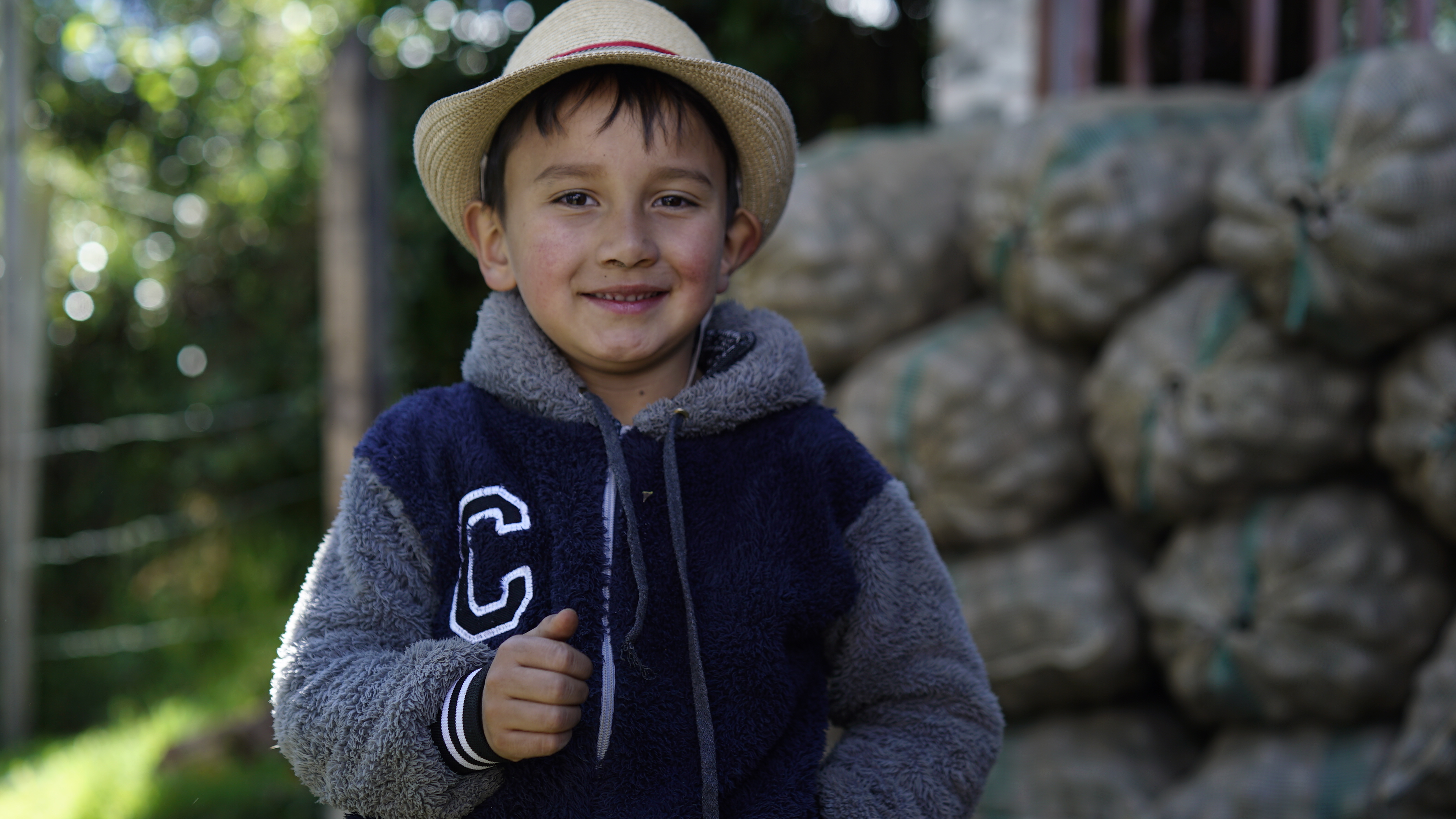 Fotografía de un niño con saco azul y sombrero sonriendo, en el fondo hay bultos de papa y el contexto es una finca productora de este alimento en Cundinamarca