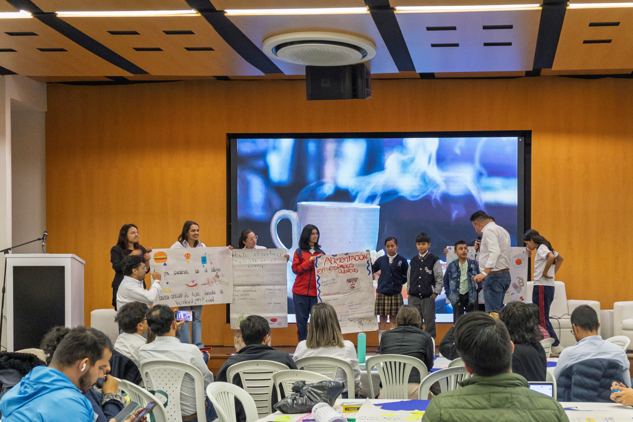 Fotografía en el auditorio de la Gobernación de Cundinamarca que muestra a niños y niñas que participaron en la mesa temática sobre seguridad alimentaria, algunos de ellos tienen uniforme de sus colegios y sostienen carteleras realizadas durante el ejercicio de participación 