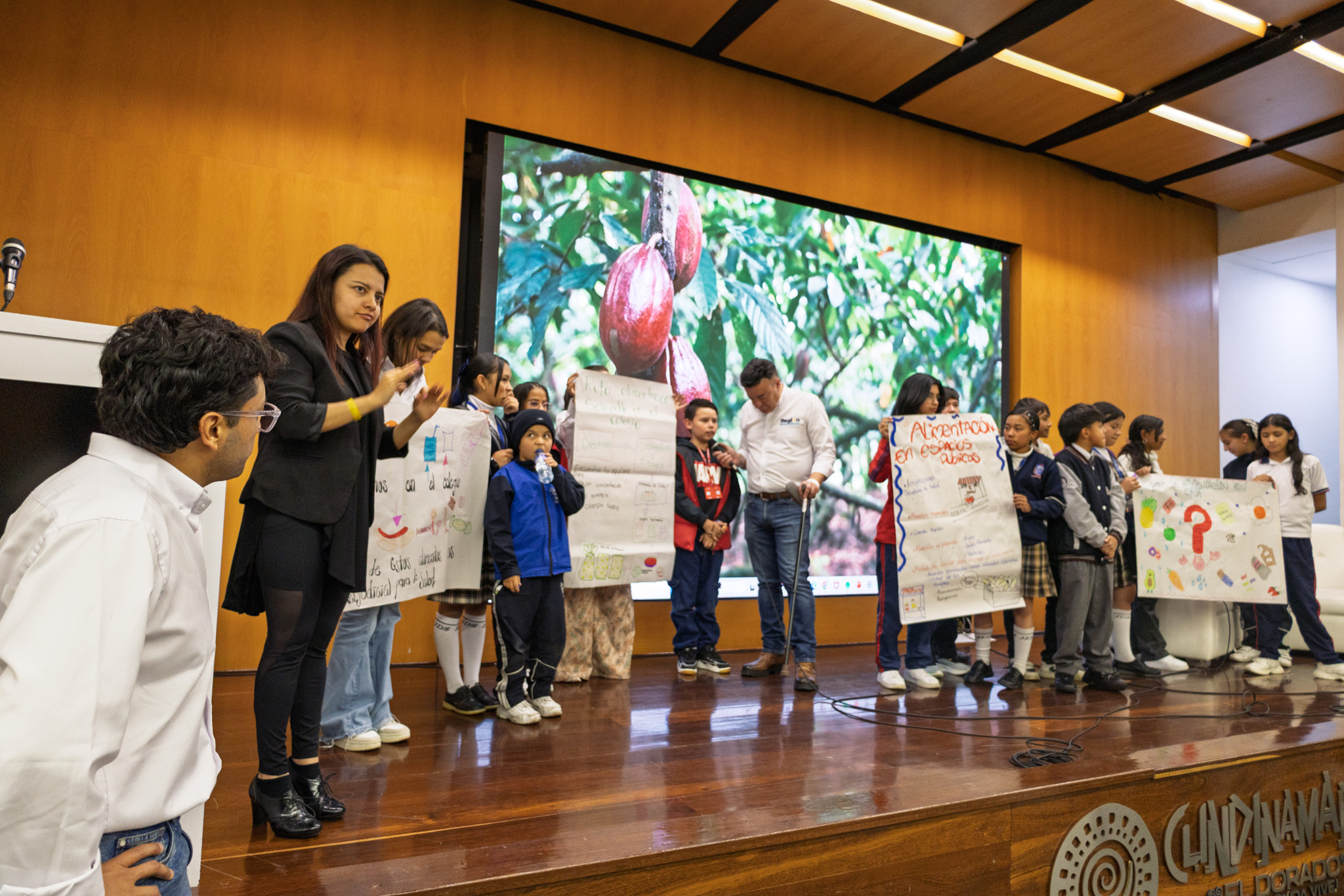 Fotografía de niños que participaron en la mesa temática sobre seguridad alimentaria, realizada en el auditorio de la Gobernación de Cundinamarca, los niños están presentado carteleras y detrás de ellos está proyectado un video