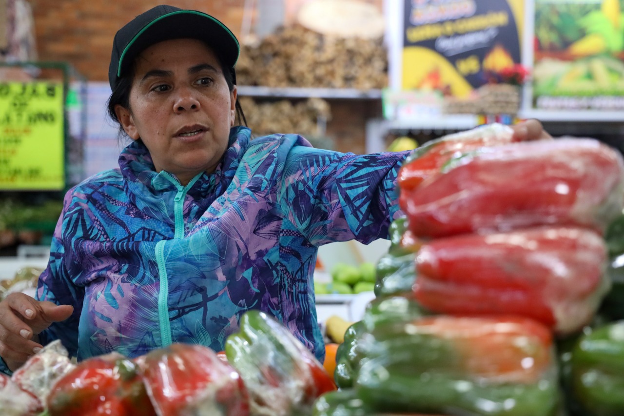 Fotografía de una mujer vestida con chaqueta de color azul y púrpura y una gorra en un mercado de víveres, varios pimentones aparecen en un primer plano difuminado