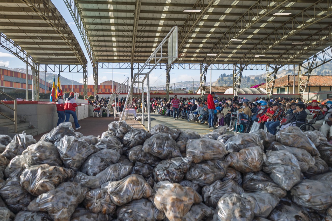 Fotografía de bultos de papas en un coliseo, en el segundo plano, aparecen muchas personas