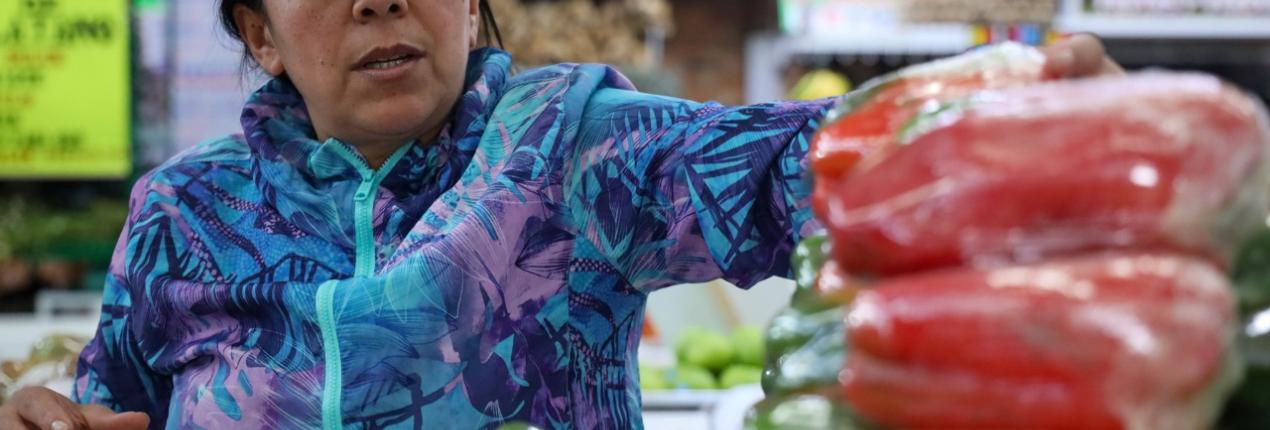 Fotografía de una mujer vestida con chaqueta de color azul y púrpura y una gorra en un mercado de víveres, varios pimentones aparecen en un primer plano difuminado