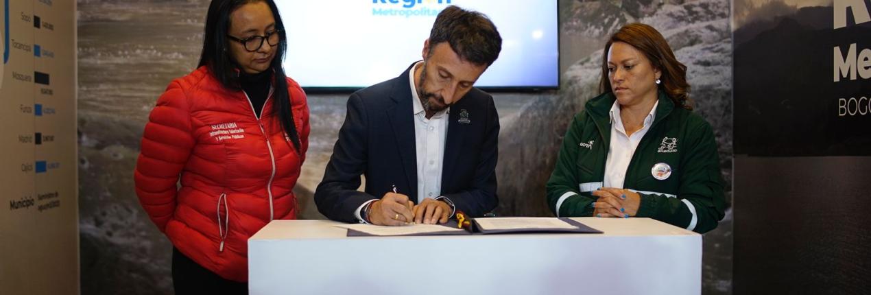 Fotografía en el que aparecen tres personas en la firma de un convenio para beneficiar a más de 40 mil personas en Soacha por medio de proyectos estratégicos en materia de acueducto y alcantarillado. El convenido fue firmado por funcionarios de la Alcaldía de Soacha, la Empresa de Acueducto y Alcantarillado de Bogotá y la Región Metropolitana Bogotá - Cundinamarca