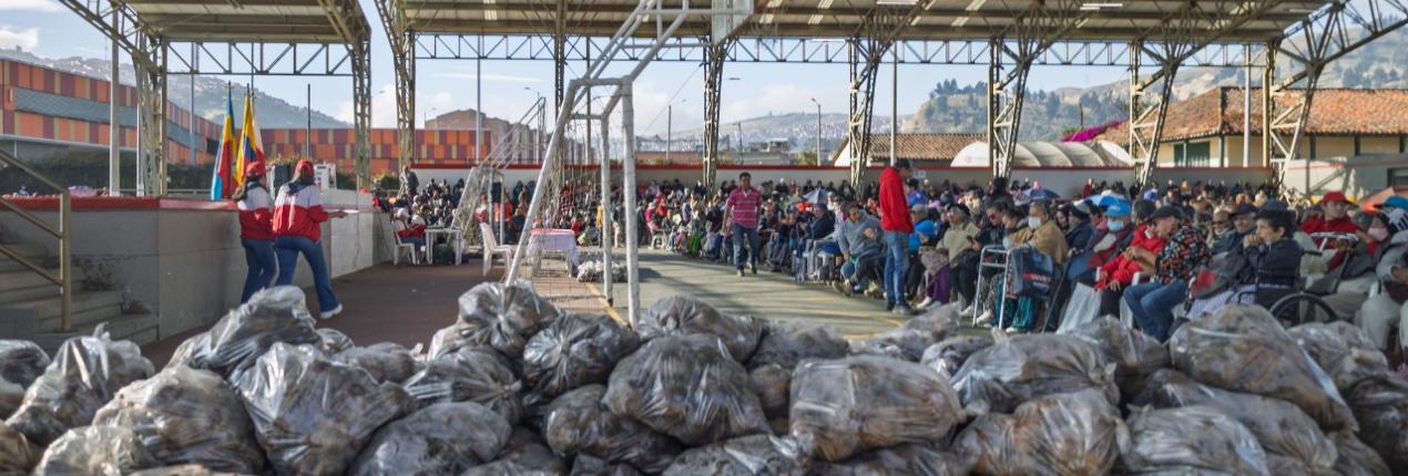 Fotografía de bultos de papas en un coliseo, en el segundo plano, aparecen muchas personas