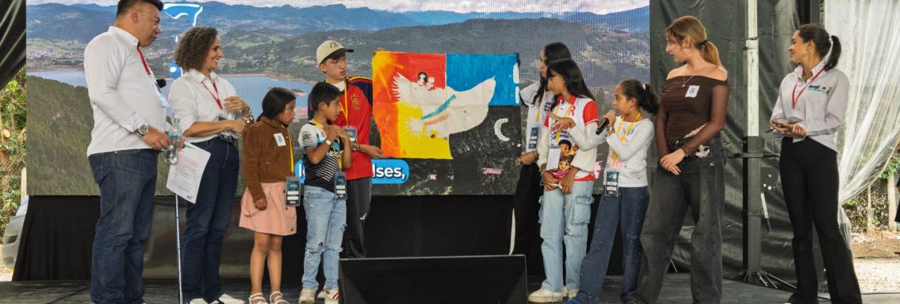Fotografía en la que aparecen siete niñas, niños y adolescentes en un escenario que tiene un fondo de pantalla, espacio en el que sostienen una bandera; los menores están acompañados de tres adultos