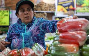 Fotografía de una mujer vestida con chaqueta de color azul y púrpura y una gorra en un mercado de víveres, varios pimentones aparecen en un primer plano difuminado