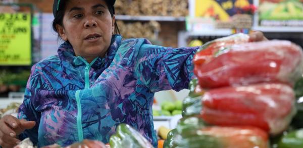 Fotografía de una mujer vestida con chaqueta de color azul y púrpura y una gorra en un mercado de víveres, varios pimentones aparecen en un primer plano difuminado